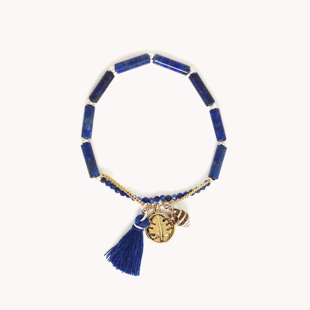 Bracelet extensible composé de perles longues et de petites perles rondes facettées en lapis lazuli, agrémenté d'une chainette dorée à l'or fin dotée d'une médaille avec un motif feuille de monstera, d'un coquillage et d'un pompon bleu à franges.