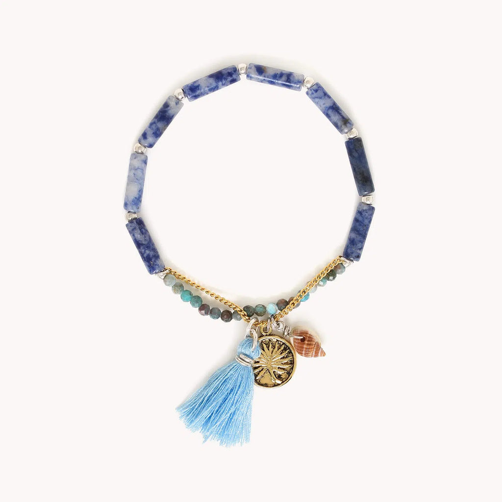 Bracelet extensible composé de perles longues, de petites perles rondes facettées en agate et d'une chainette dorée à l'or fin dotée d'une médaille avec un motif feuille de palme, d'un coquillage et d'un pompon bleu à franges.